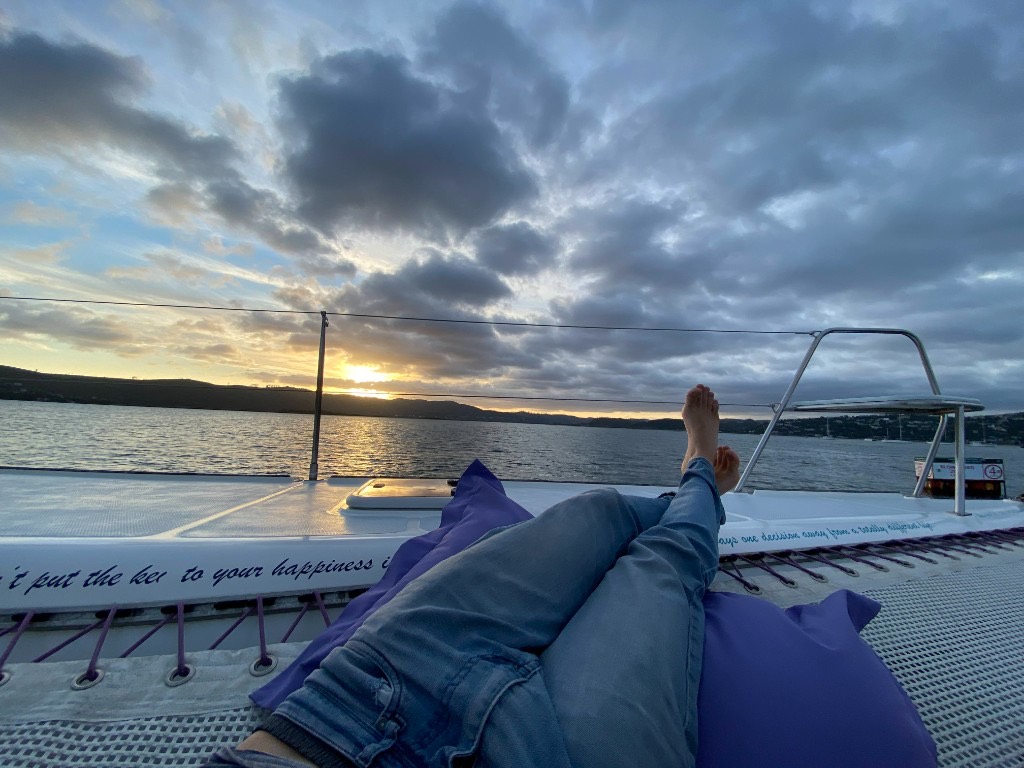 Bare feet on a purple beanbag on a catamaran trampoline at sunset, with the Knysna lagoon and a low orange sun ahead.