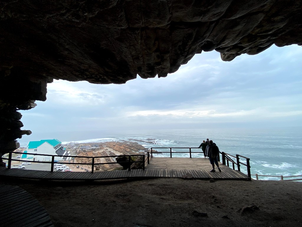 Looking out from the dark mouth of Pinnacle Point cave onto a wooden viewing deck, the rocky Mossel Bay coastline, and a grey sea.
