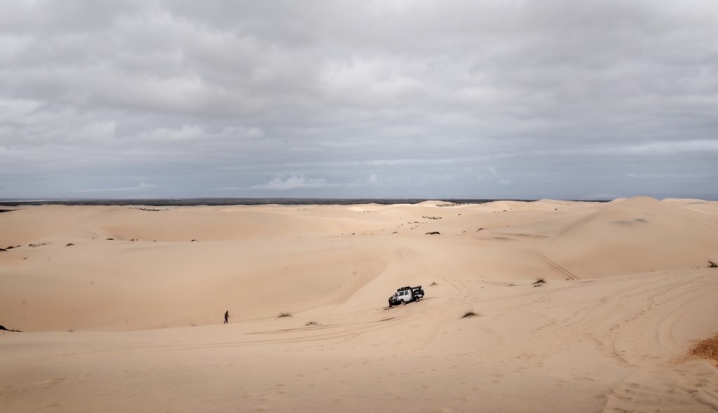A white bakkie parked on pale sand dunes under an overcast sky, with tyre tracks and a person walking on the dune.