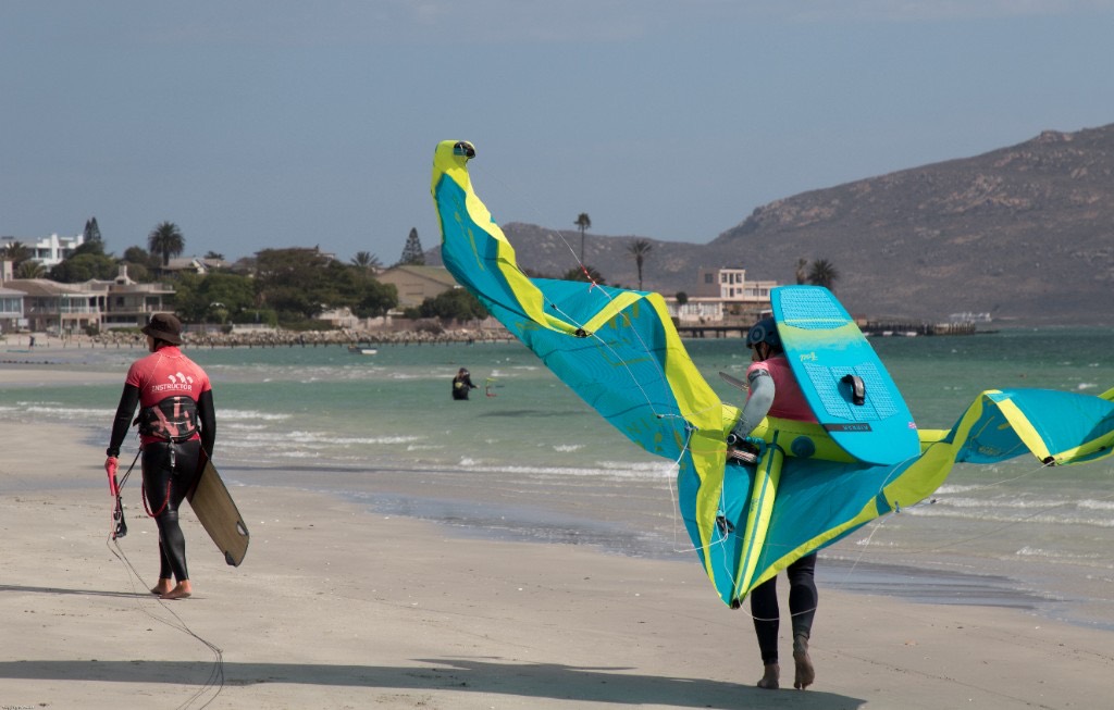 Two kitesurfers walking on Langebaan lagoon sand; one carries a blue and neon kite and a turquoise board, the other wears a red instructor shirt.