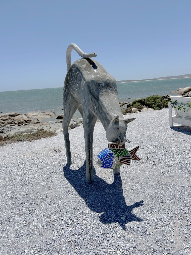 A spindly grey cat sculpture holding a mosaic fish in its mouth, standing on pebbles beside turquoise water.