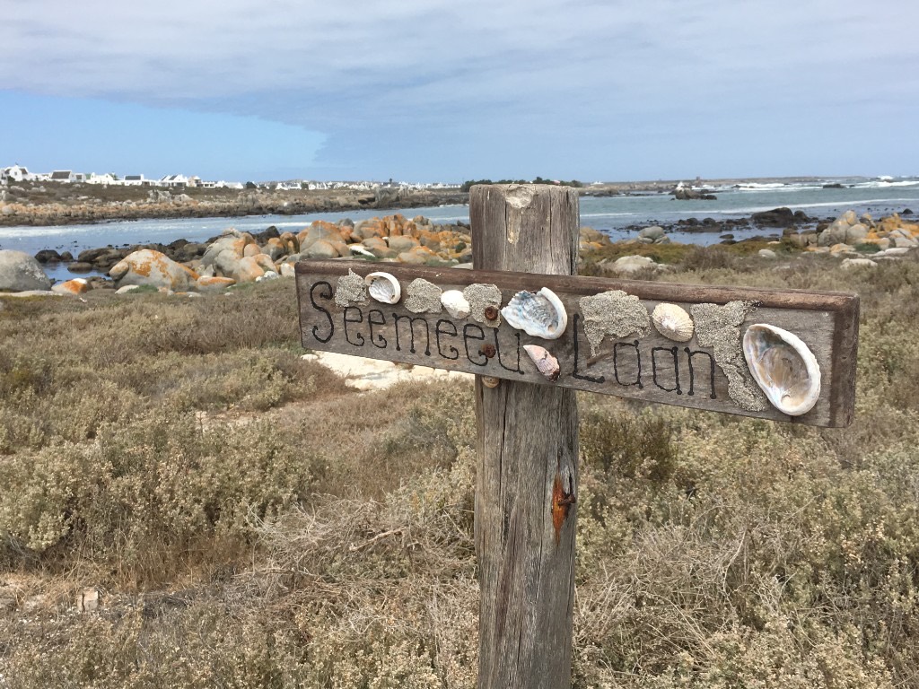 A weathered wooden Seemeeu Laan sign decorated with shells, with granite boulders and a whitewashed village across the water.