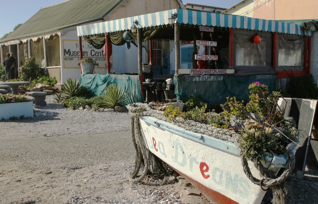 A white wooden boat planter painted Sea Dreams, with a snack bar and the Port Nolloth Museum behind it.