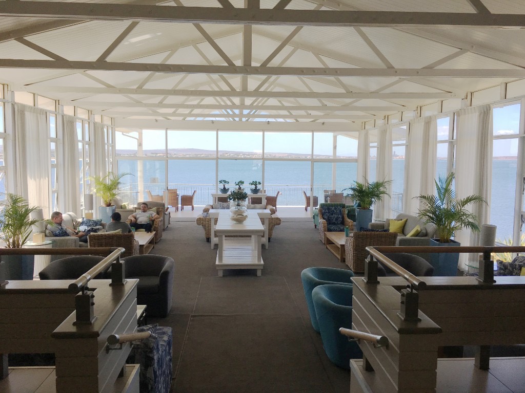 A bright coastal lounge with white truss ceilings and floor-to-ceiling windows overlooking the bay.